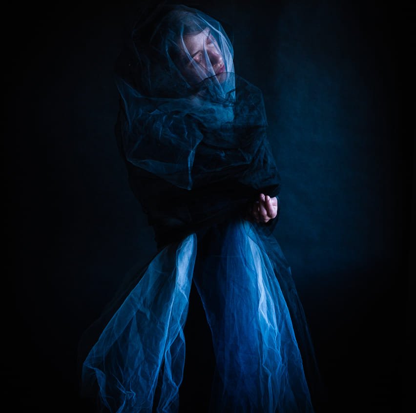 A figure is draped in gauzy fabric on a moody, blue-lit stage. Her eyes are closed and her face is shrouded by a thin veil.