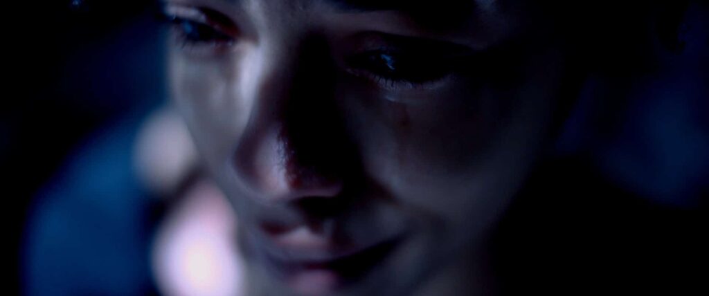 Closeup of a woman’s face, in a dark space with small dapples of light on her nose and cheeks. She arrears to be crying.