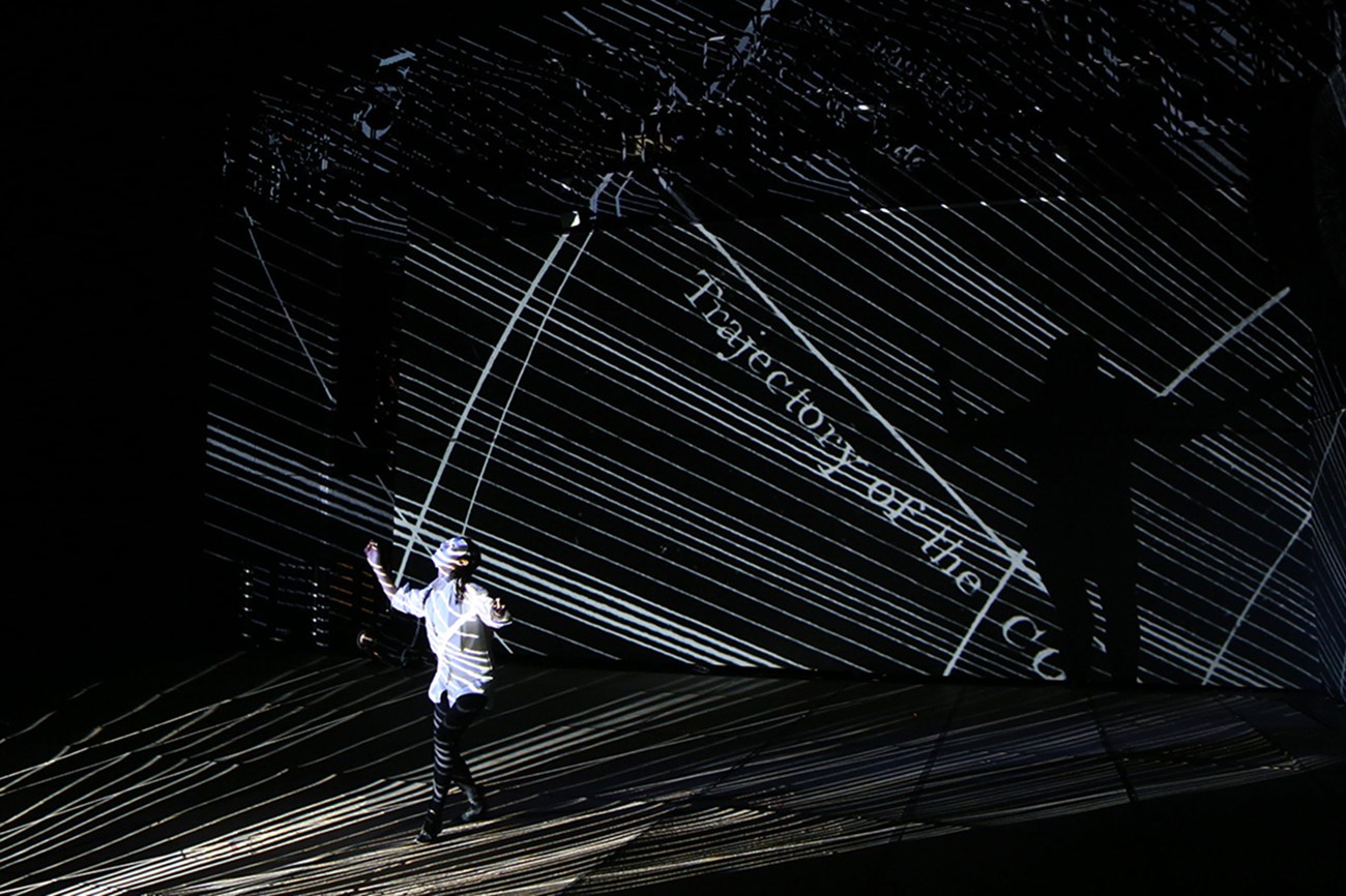 A person stands on a dark stage, the whole stage is covered with a projection of diagonal white lines. Some text in the background reads, “Trajectory of the C…”