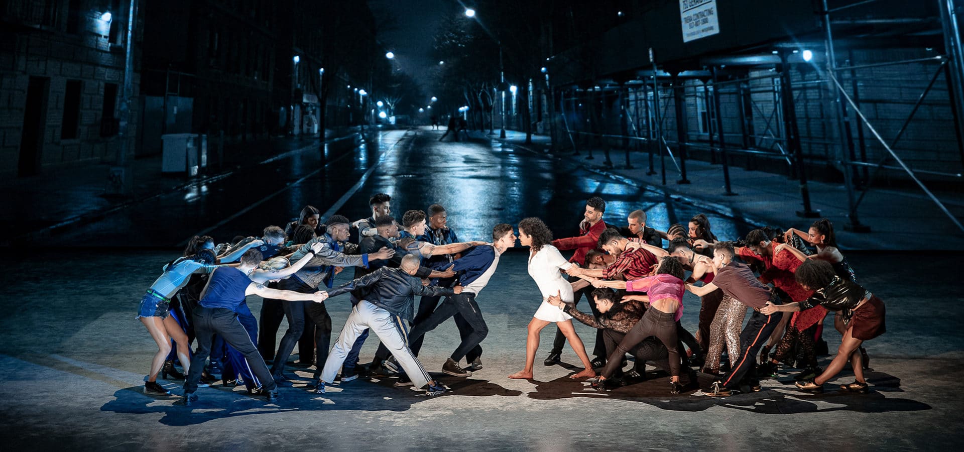 Two groups of dancers face each other, pulling the two characters in the center in opposite directions. The left group wears shades of blue and the right group wears shades of red. They are on a dark city street. 