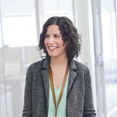 A woman with curly hair and a gray blazer smiles in an office setting.