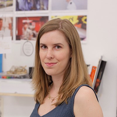 Woman with shoulder-length brown hair in a blue sleeveless top, standing indoors with blurred photos and books in the background.