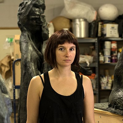 Person in an art studio with large sculptures and art supplies in the background.