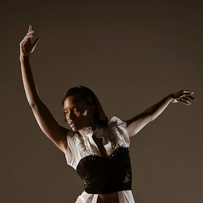 Person in a graceful dance pose with one arm raised, wearing a white top and black corset, against a brown background.