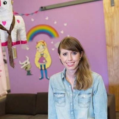 Woman in a denim shirt in front of a purple wall with cartoon decorations and a unicorn piñata hanging nearby.