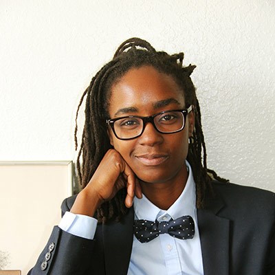 Person with dreadlocks and glasses wearing a navy suit and polka dot bow tie.