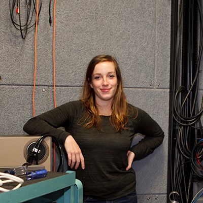 Woman standing in a workshop with technical equipment and cables in the background.