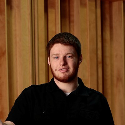 A young man with auburn hair and a beard, wearing a black shirt, stands in front of a wooden panel background.