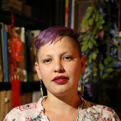 Person with short purple hair and floral shirt in a room with books and decorations in the background.