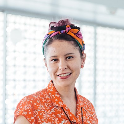 Person smiling indoors with a colorful headscarf and floral shirt.