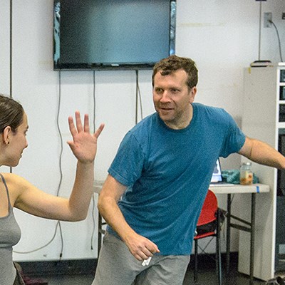 A man and woman engaged in a dance rehearsal in a studio, with a television and furniture in the background.