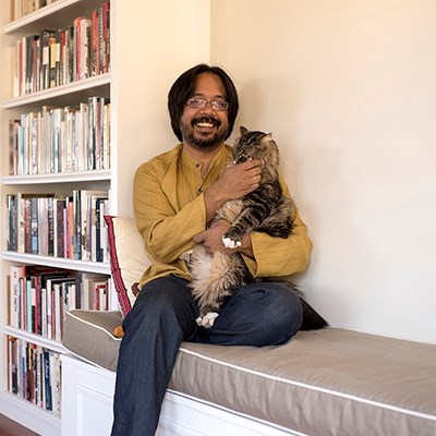 Person sitting on a cushioned bench holding a large fluffy cat, with a bookshelf in the background.