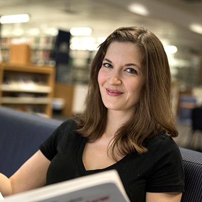 Person sitting in a library, holding a book and smiling at the camera.