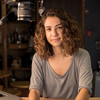 A person with wavy brown hair in a gray shirt sits at a table with photography equipment overhead.