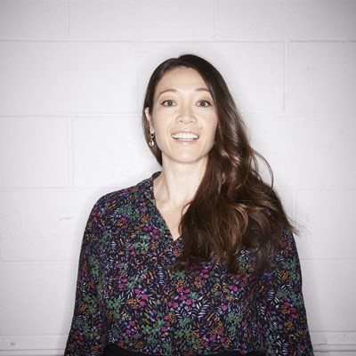 Person smiling in a floral blouse against a gray brick wall.