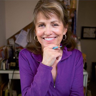 A person with light brown hair, wearing a purple top, smiles and holds a pen against their chin in a home office setting.