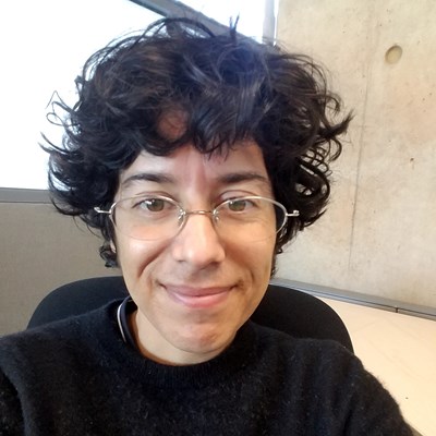 Person with curly hair and glasses smiling, sitting in front of a concrete wall and window.