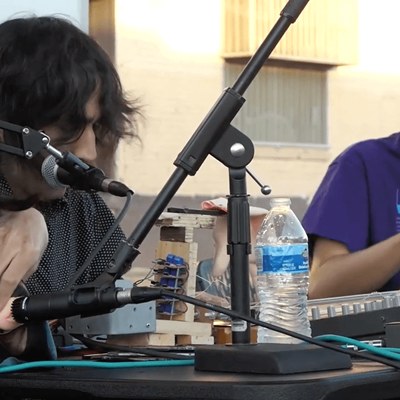 Two people seated at a table with electronic equipment and a microphone setup outdoors.