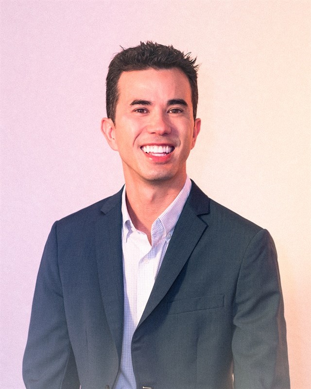 A man in a dark blazer and light shirt smiling against a warm gradient background.