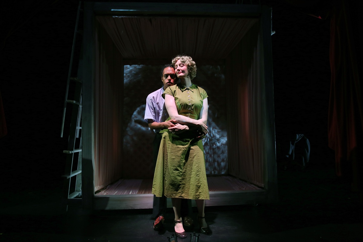 Two people stand closely together on a dimly lit stage, embraced within a box-like structure.