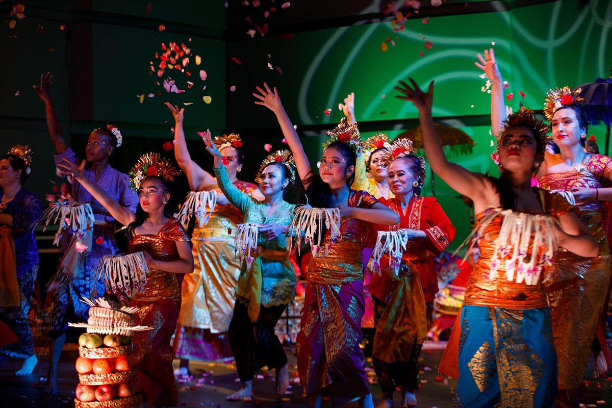 Dancers in colorful traditional Balinese costumes perform on stage, scattering flower petals.