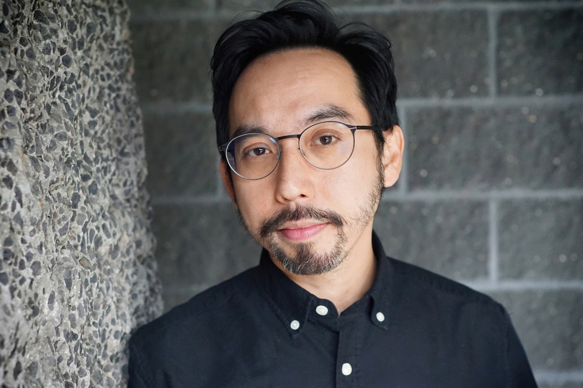 A person with glasses and a black shirt stands against a textured wall and brick background.