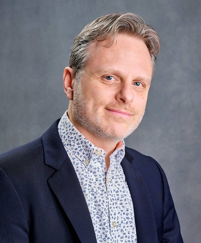 Man in a navy blazer with a white floral shirt against a gray background.