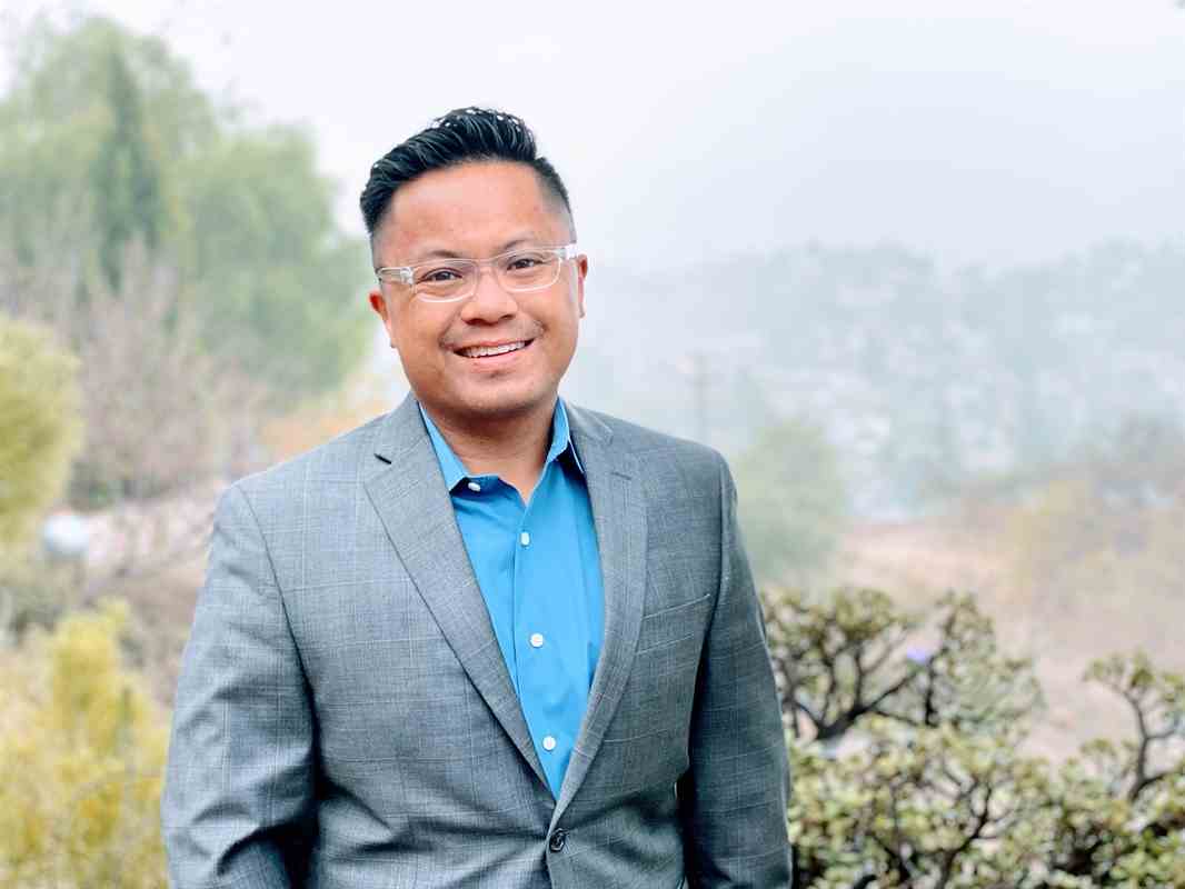 Man in a gray suit and blue shirt standing outdoors with trees and shrubs in the background.