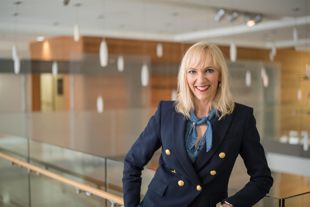 Smiling woman in a navy blazer with gold buttons in an office setting.