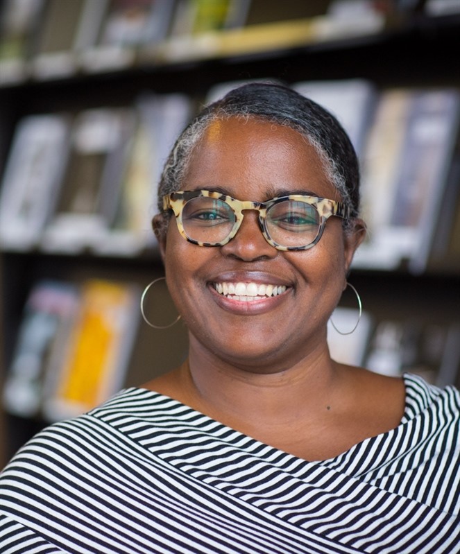 Smiling person with glasses and striped outfit in front of blurred shelves.