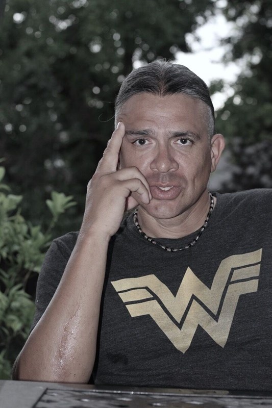 Person sitting outdoors with greenery in the background, wearing a dark t-shirt with a yellow "W" logo.