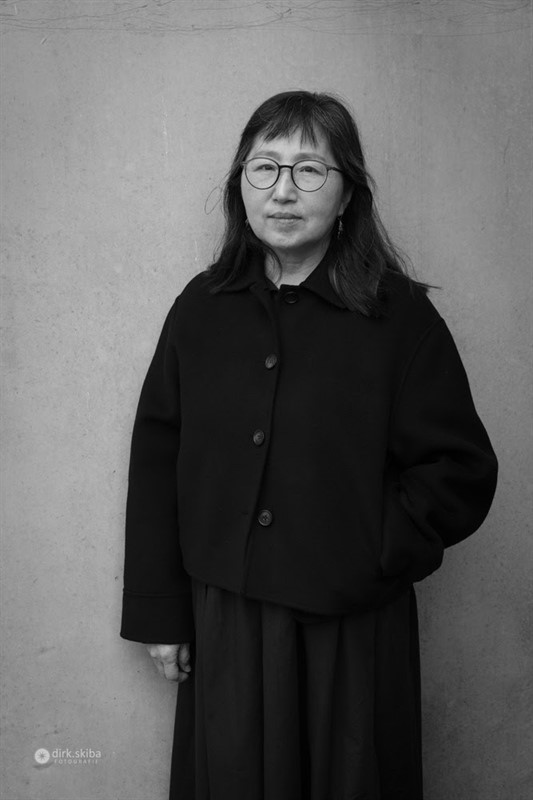 Person in a dark coat standing against a light wall.