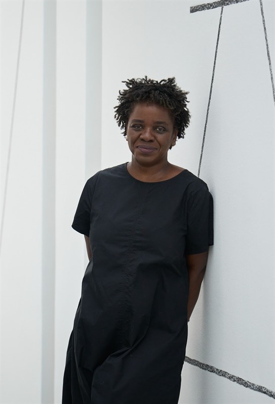 Person standing against a white wall with geometric line designs, wearing a black dress.