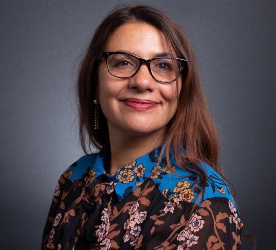 Portrait of a smiling person with glasses and a floral blouse against a grey background.