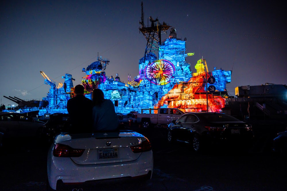 Large ship with colorful light projections at night, with silhouettes of two people sitting on a car in the foreground.