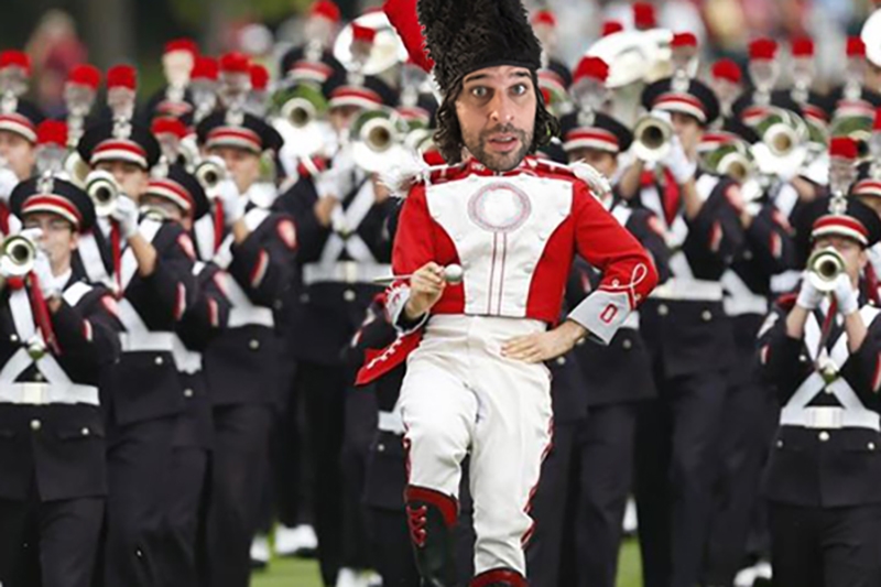 A marching band leader in a red, white, and black uniform holding a baton, with band members in similar uniforms playing brass instruments in the background.