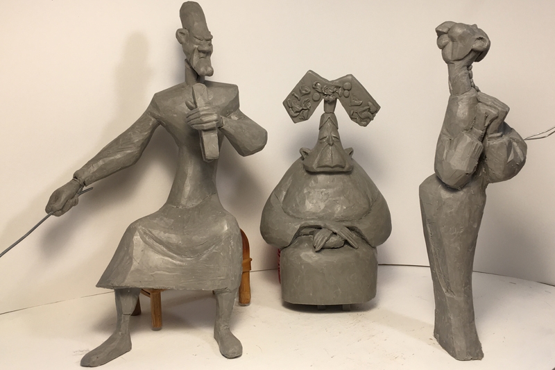 Three stylized gray sculptures: a seated figure with a rod (left), a rotund seated figure with an elaborate headpiece (center), and a standing figure with a string instrument (right).