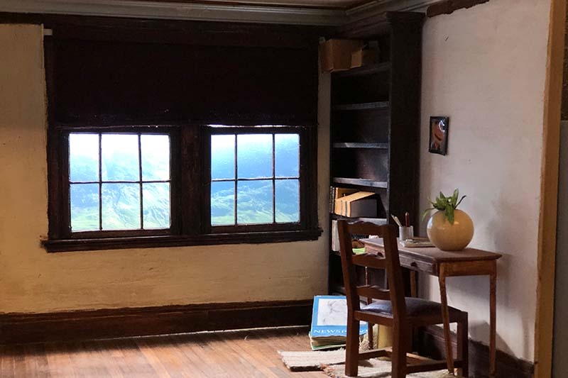 A rustic room with a window showing mountains, a desk with a vase and pens, and an empty bookshelf.