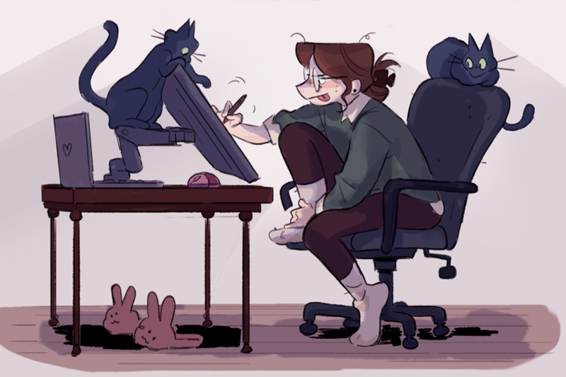 Cartoon of a person working at a desk with a drawing tablet, surrounded by two black cats and a pair of pink bunny slippers on the floor.