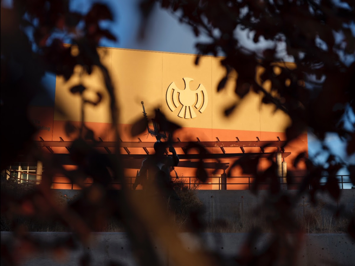 A beige building facade with a stylized bird emblem, partially obscured by dark tree branches and leaves.