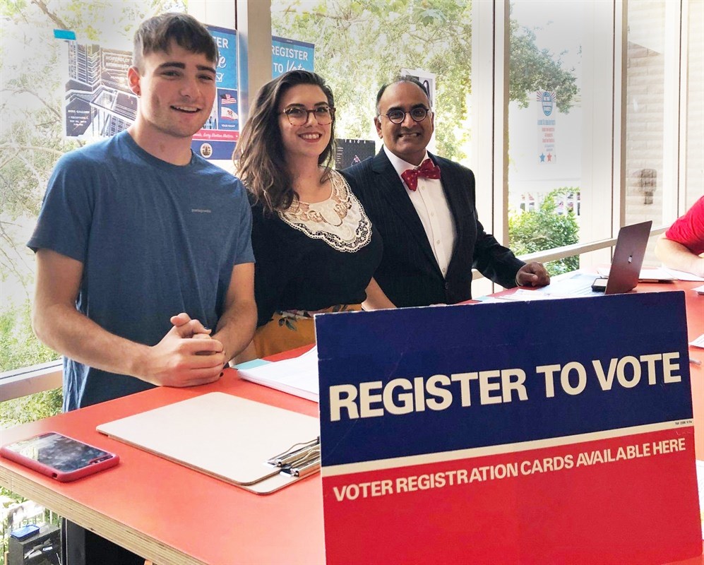 Three people standing behind a voter registration table with a "REGISTER TO VOTE" sign.