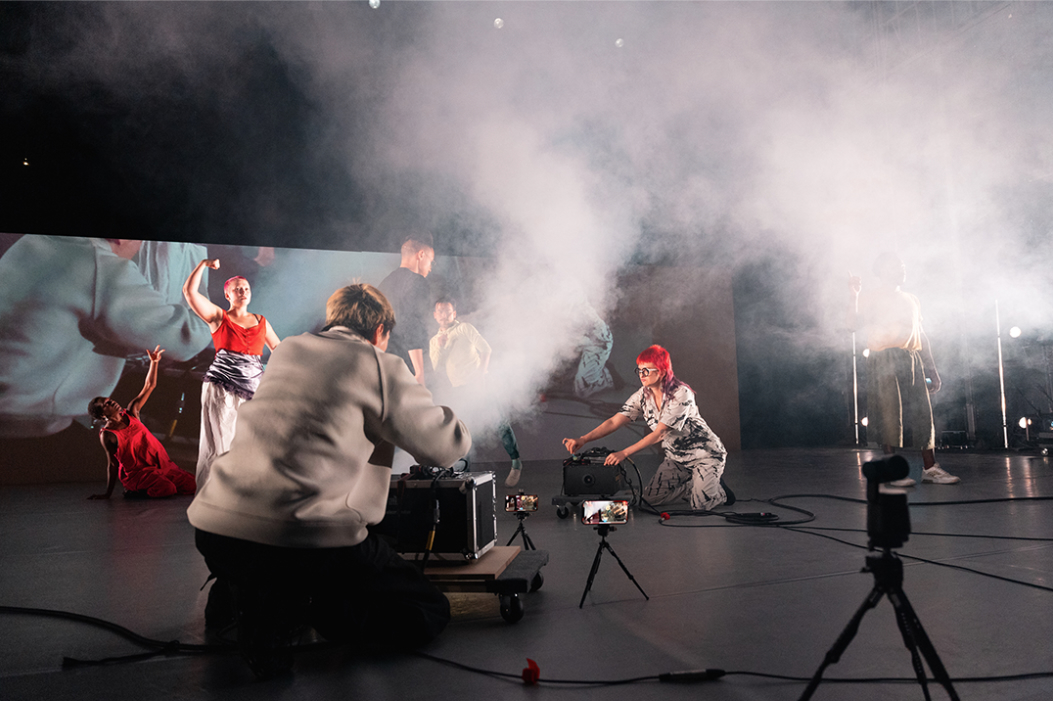 People work with audio and video equipment on a stage, as other people dance. Smoke swirls around in the air.