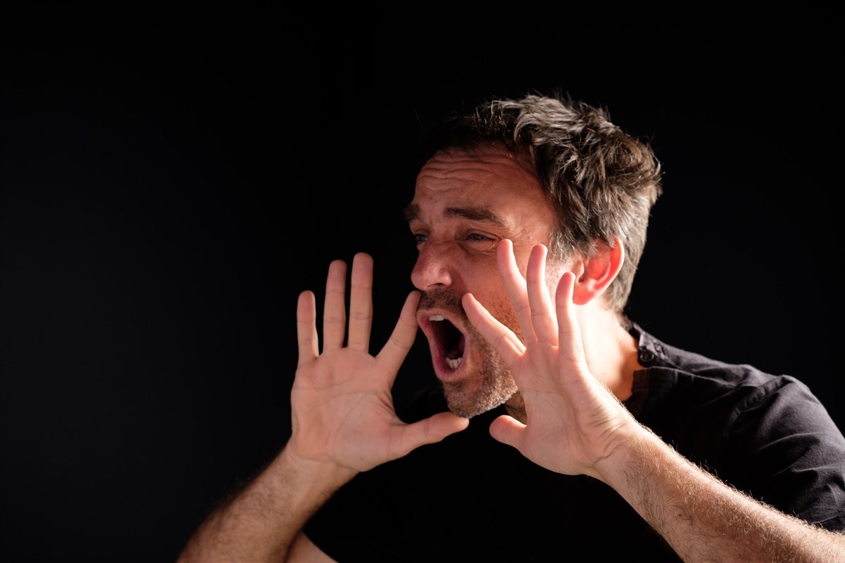 A person with raised hands and an open mouth, possibly shouting, against a dark background.