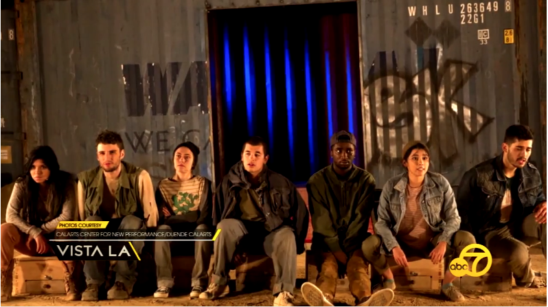 Seven people sitting on crates in front of a blue shipping container with graffiti.