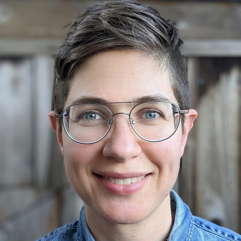 Close-up portrait of a person wearing glasses and a denim shirt, with a wooden background.