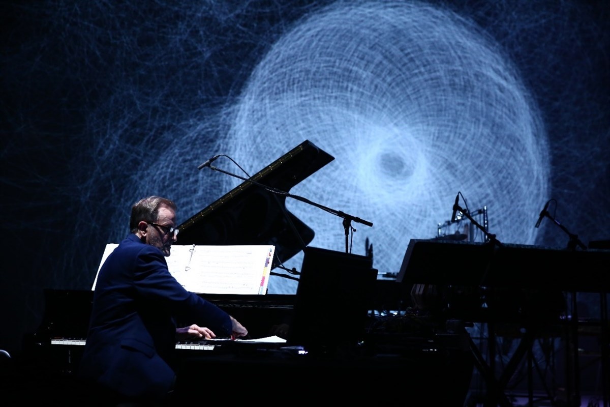 Person playing a grand piano in front of a swirling abstract projection.