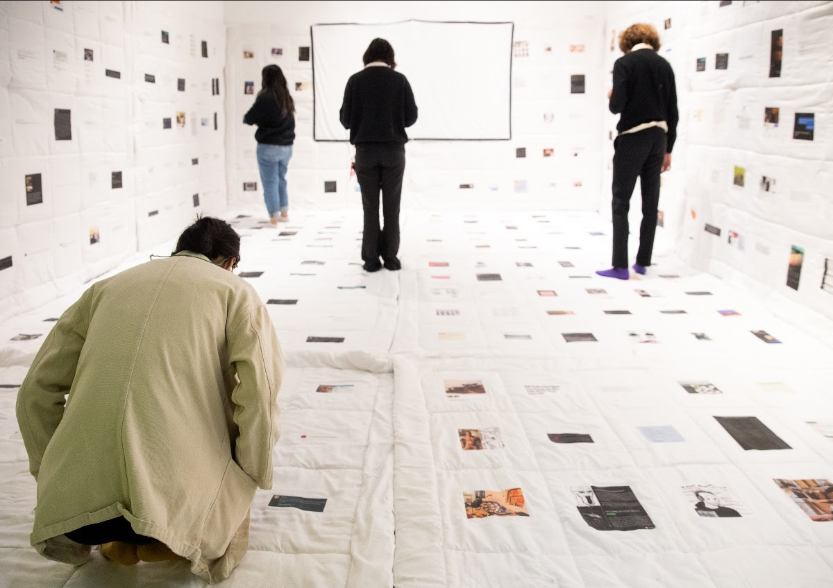 People in a white-walled room with quilt-like material featuring scattered images and text.