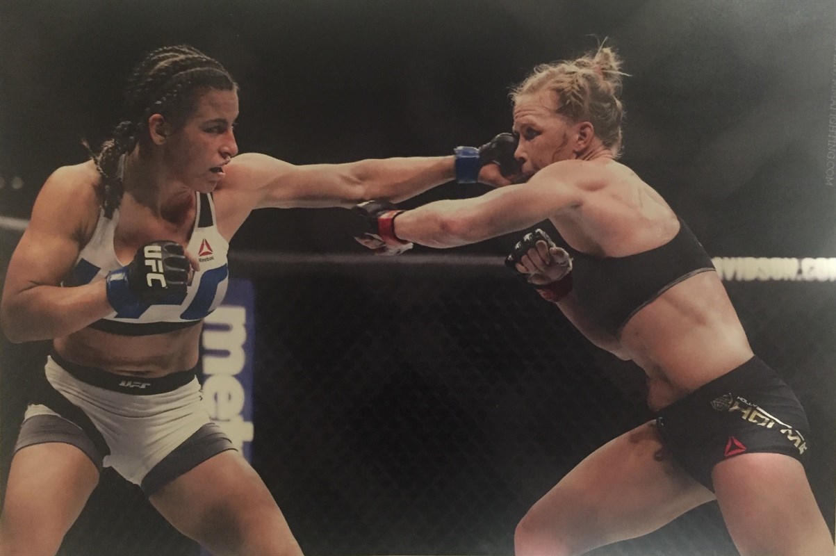 Two female mixed martial artists in a cage fight, one throwing a punch and the other blocking.