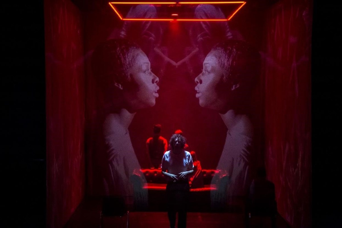 A dramatic red-lit scene with two large projected profile images facing each other and a person standing in front of a red couch.