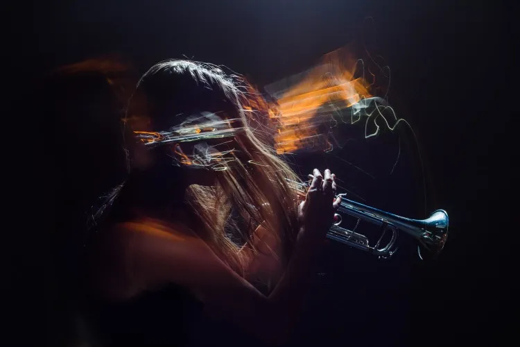 A motion-blurred photograph of a person with long hair playing a trumpet against a dark background